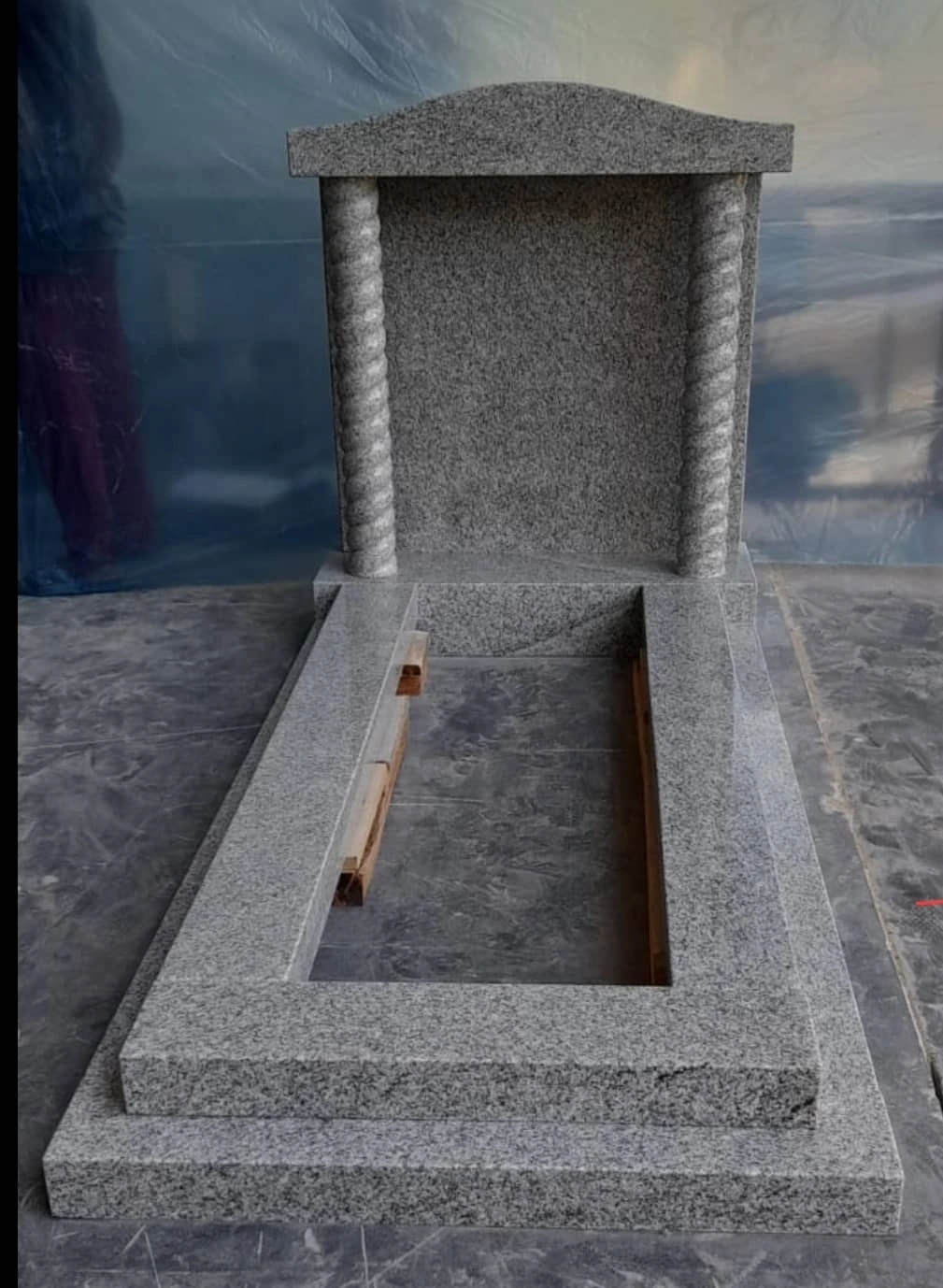 Grey memorial with a capped headstone, column details and a straight kerb surround, shown in the workshop.