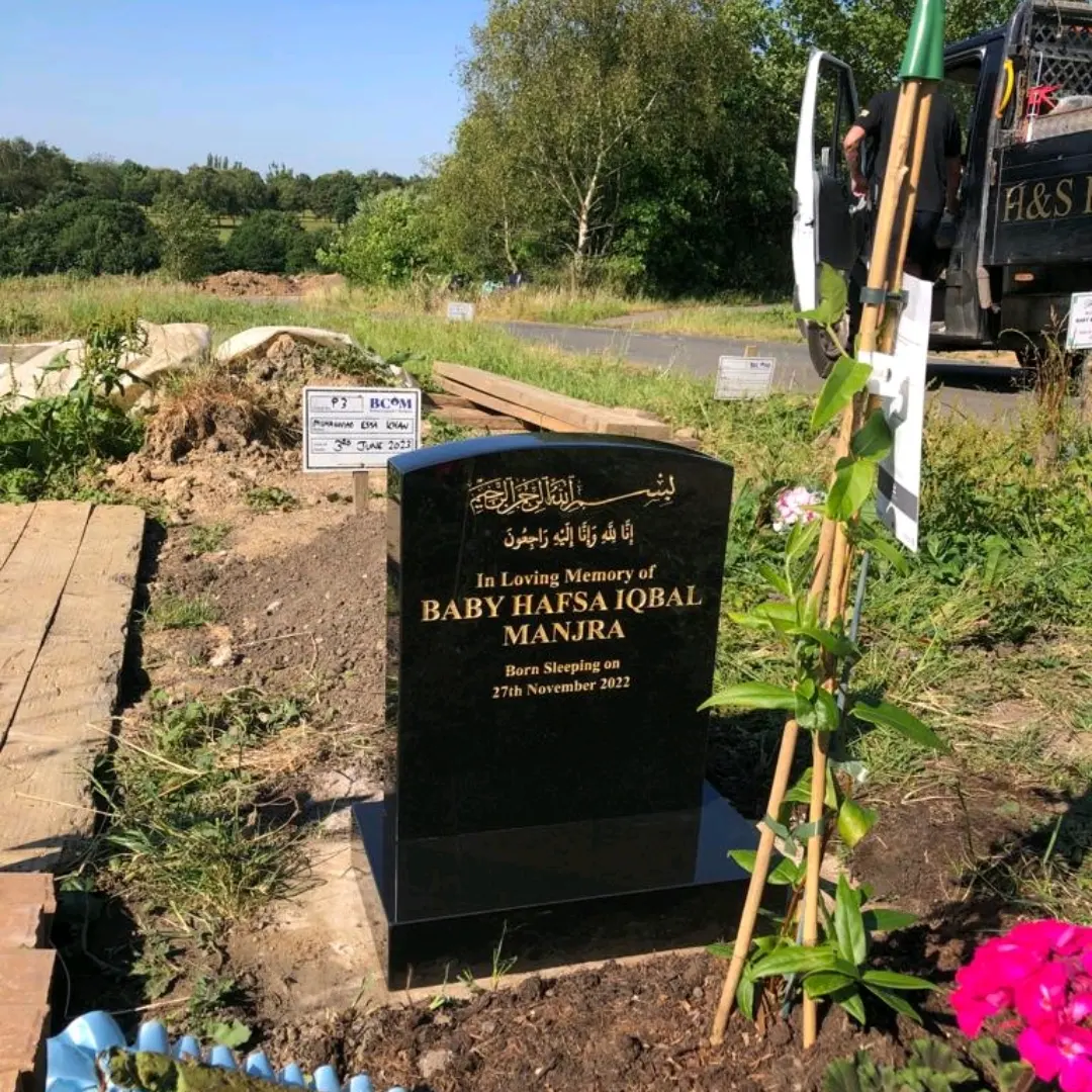 Small black headstone with a curved top, gold Arabic and English inscription, and a matching base